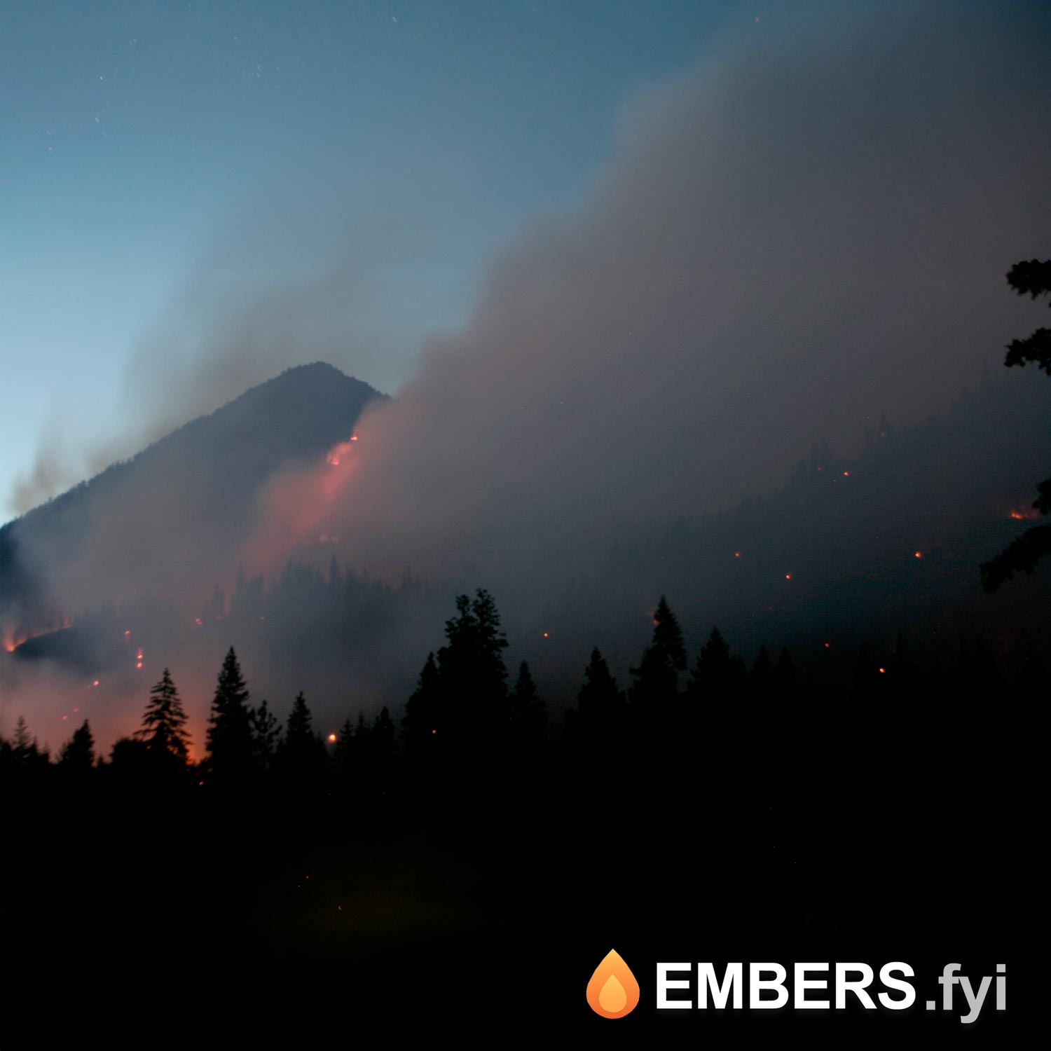 Glowing embers carried by wind above a mountainside fire at dusk